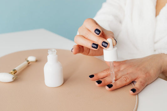 A person applying cuticle oil with a dropper beside dark-polished fingernails