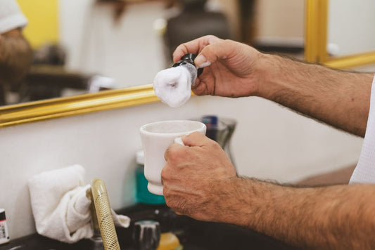 Hands holding a shaving brush covered in lather above a white shaving mug
